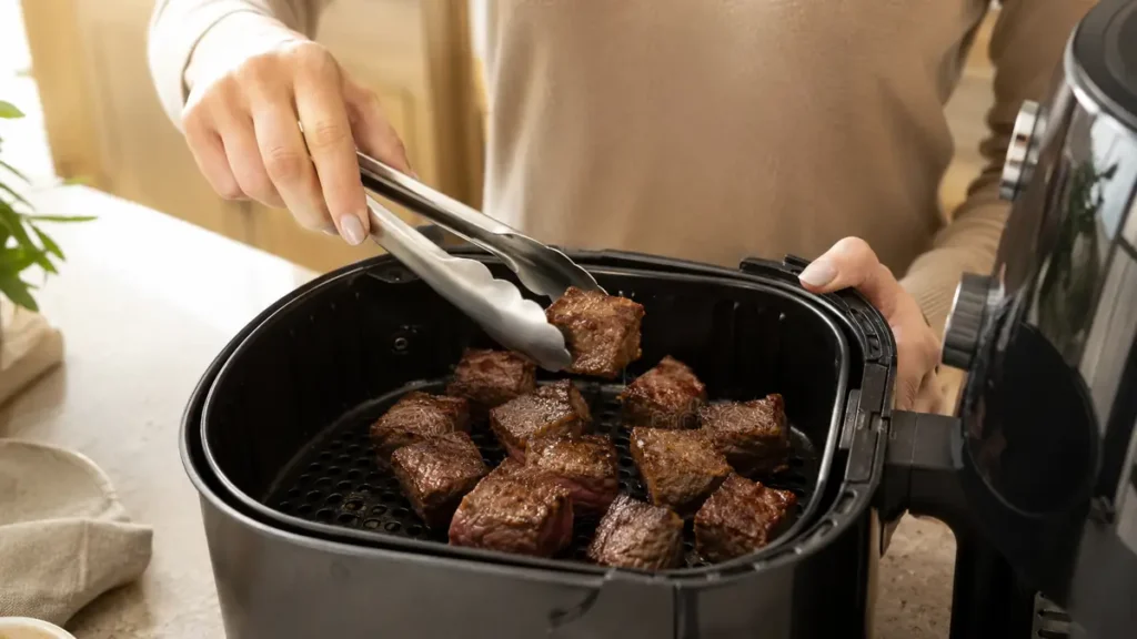 Person using tongs to lift cooked beef chunks from air fryer.